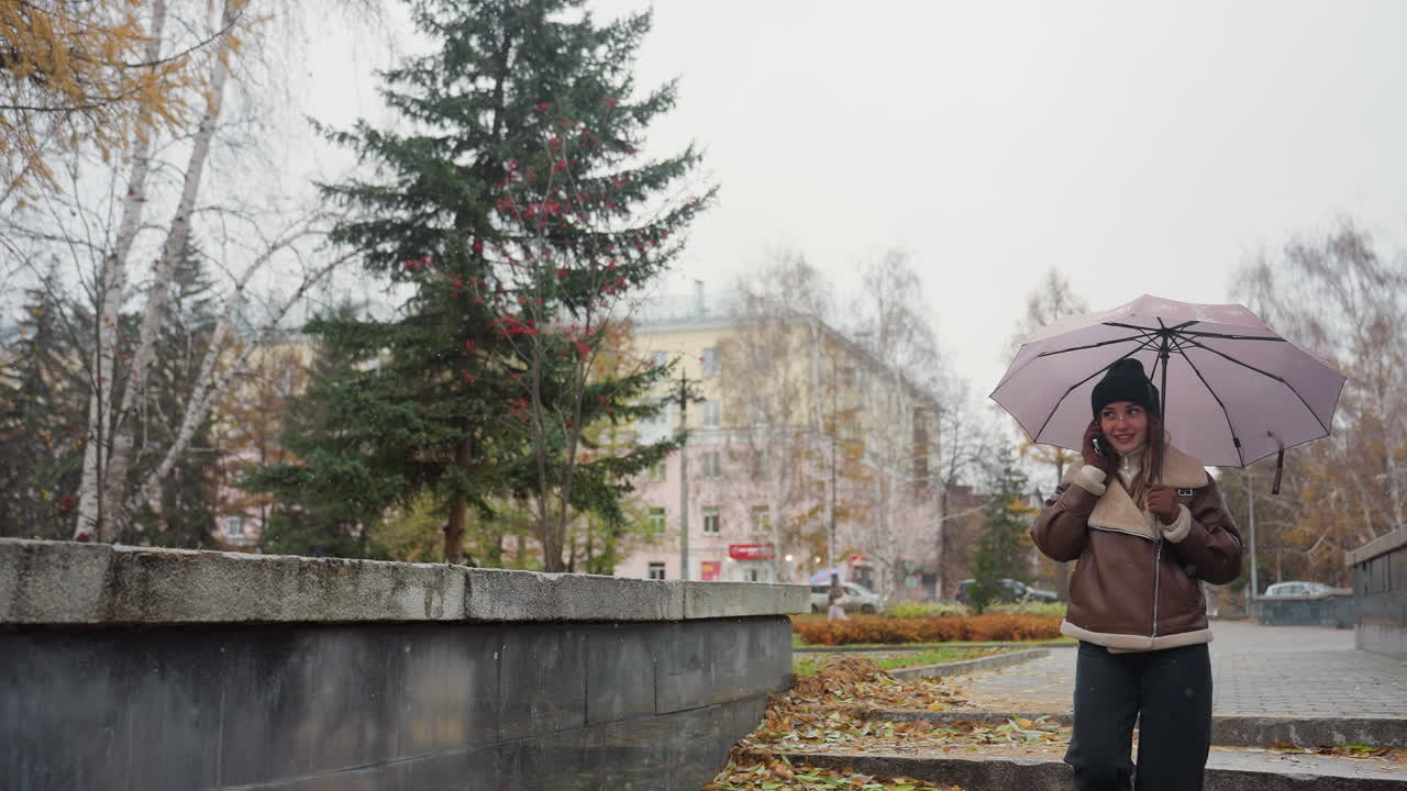 Cheerful girl smiling while making phone call, holding umbrella, wearing black knit cap, brown shearling jacket, black trousers, walking through urban park on cold overcast day with light snowfall