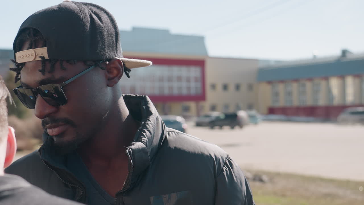 Close up of man with dark skin interacting with friend in urban street on sunny day, dressed in black puffer jacket, looking down with blurred buildings and parked cars in soft background