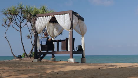 Frau-Sitzt-In-Cabana-Mit-Blick-Auf-Den-Ozean,-Umgeben-Von-Sand-Und-Palmen