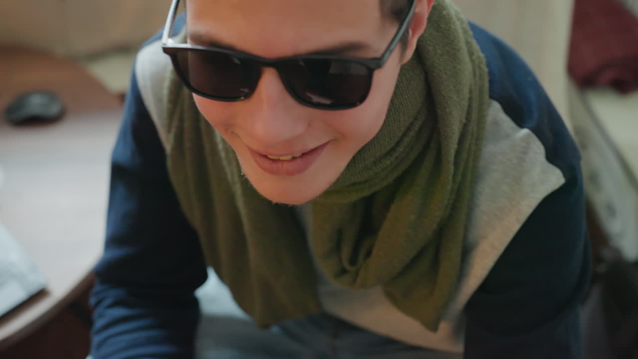 Thoughtful man adjusts sunglasses beside window with olive scarf and illustrated bookshelf, soft ambient light creating calm mood, closeup portrait capturing quiet reflection and subtle smile