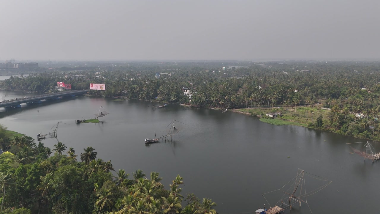 Drone footage reveals lush tropical town with dense coconut palm groves surrounding calm river waters. Colorful residential buildings with red and blue rooftops nestle among greenery. Bridge crosses w
