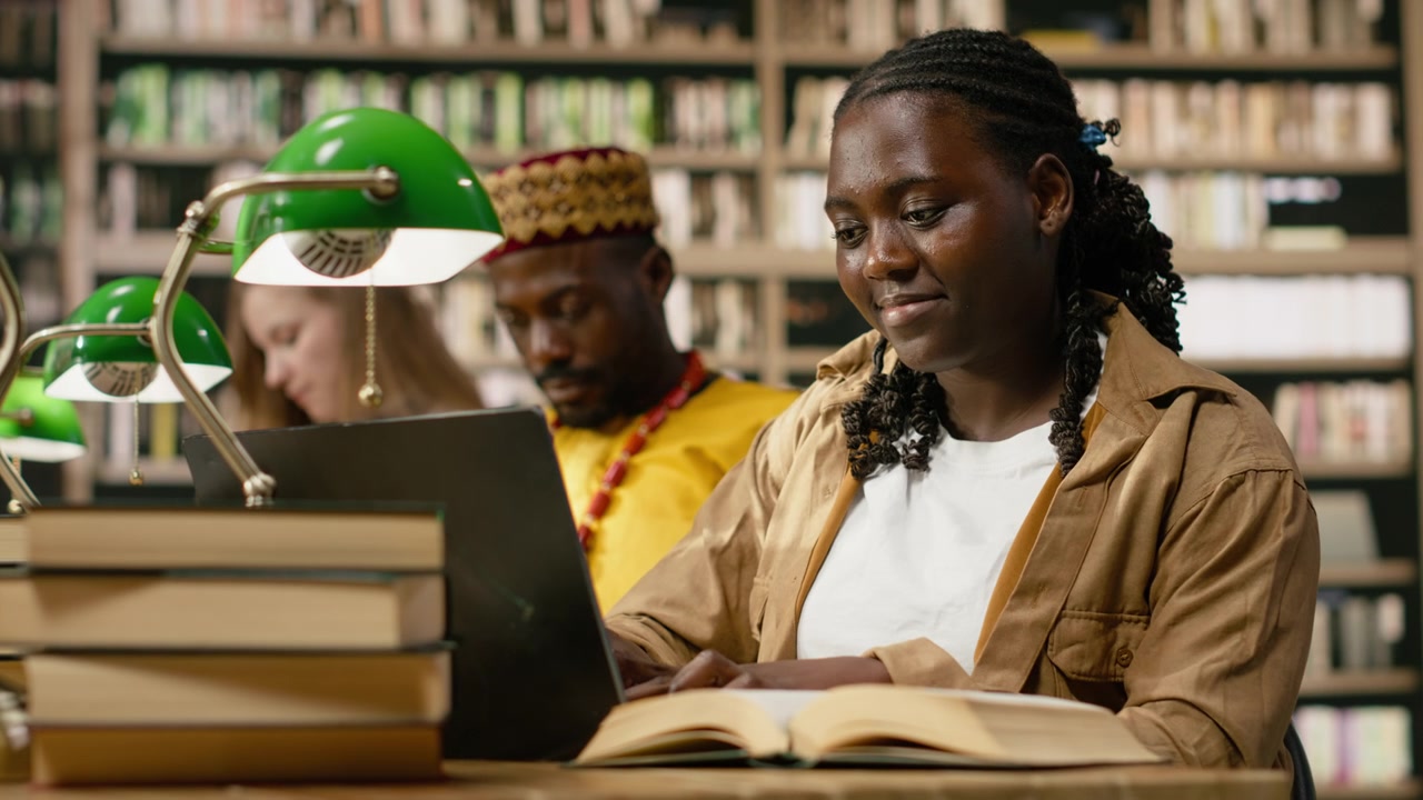 African american student typing on laptop beside open book in library space