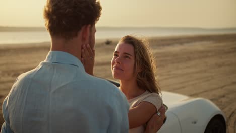 happy girl strokes the cheek of her bearded boyfriend near a white car outside. a girl and a boy on a date on a sunny windy summer evening