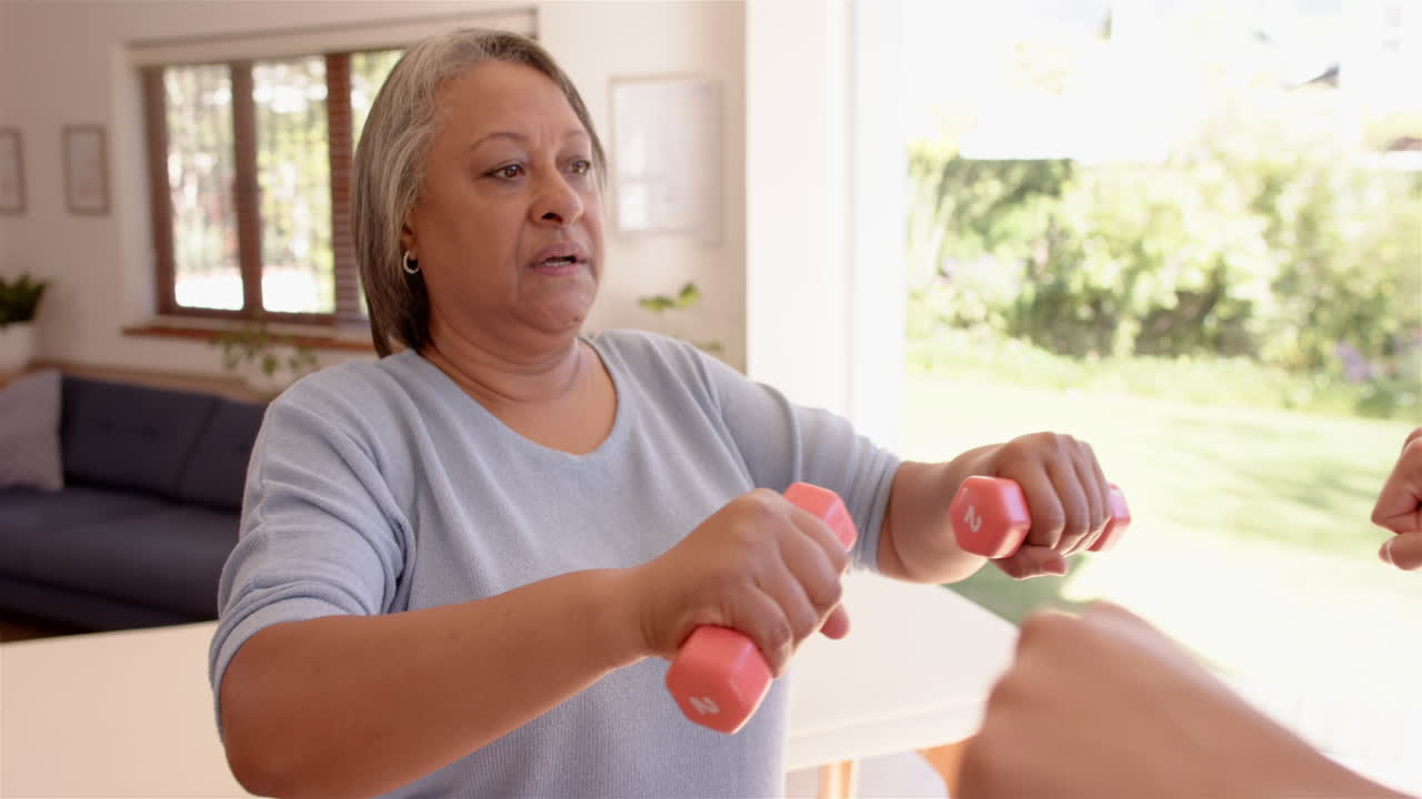 Exercising with dumbbells, senior woman guided by male physiotherapist in living room