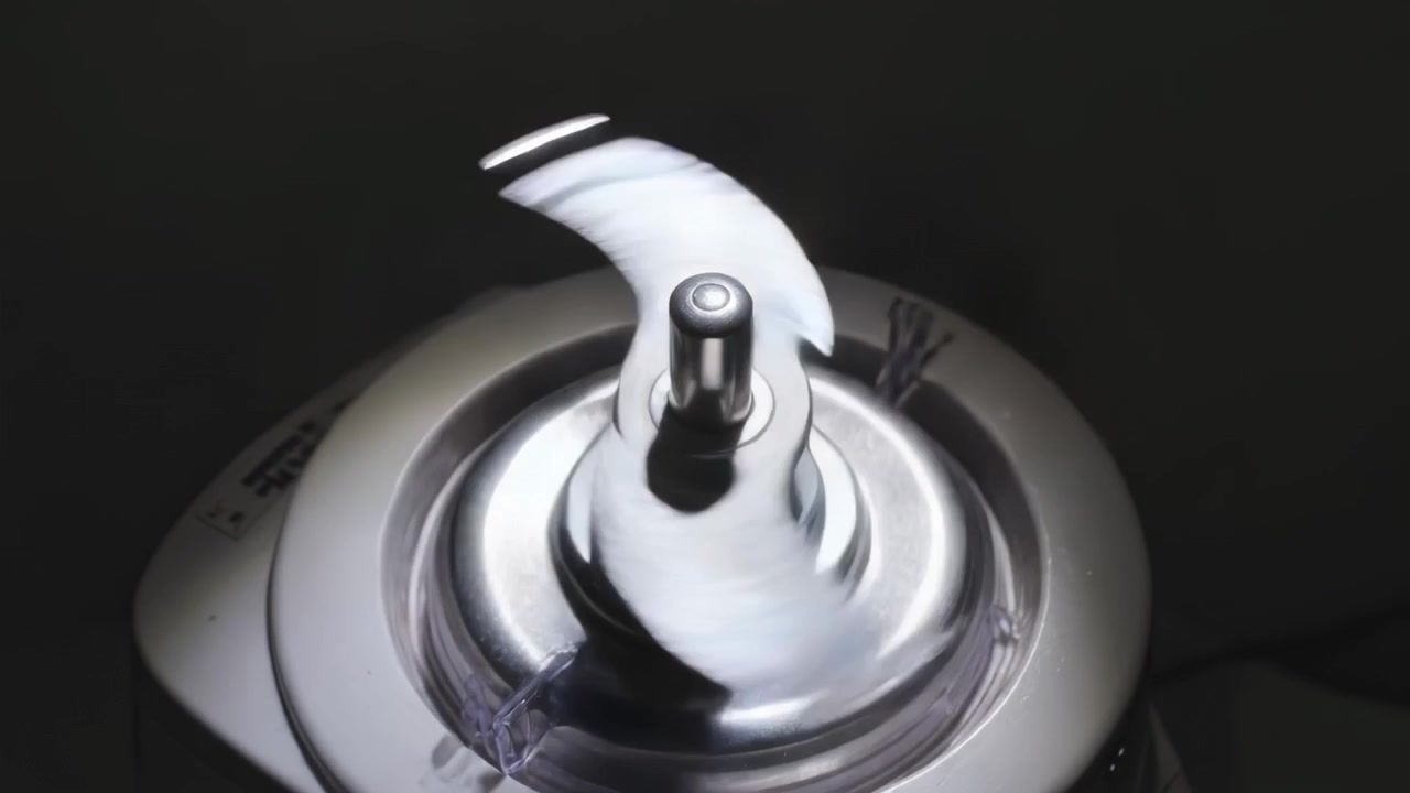 Conjunto de lâminas giratórias do liquidificador sob tampa transparente, cabo de alimentação energizando o motor misturando resíduo pálido.