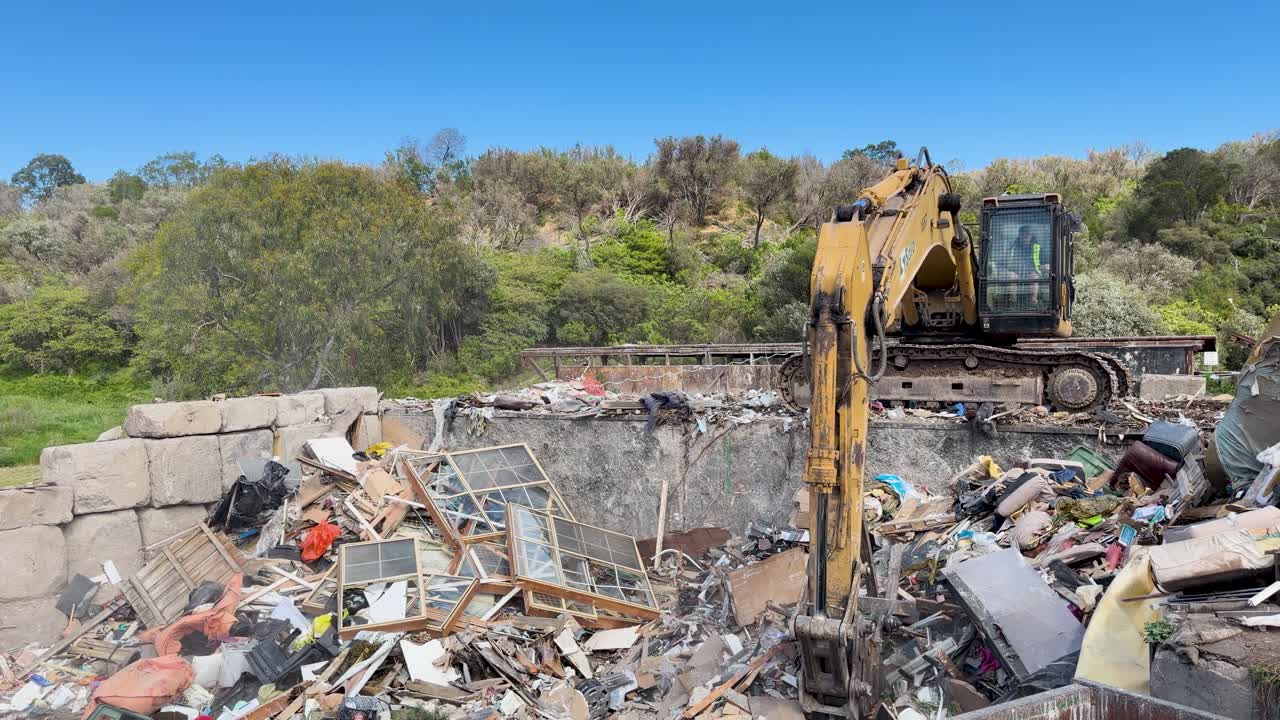 Excavator moves and sorts mixed waste in an outdoor landfill surrounded by greenery and trees