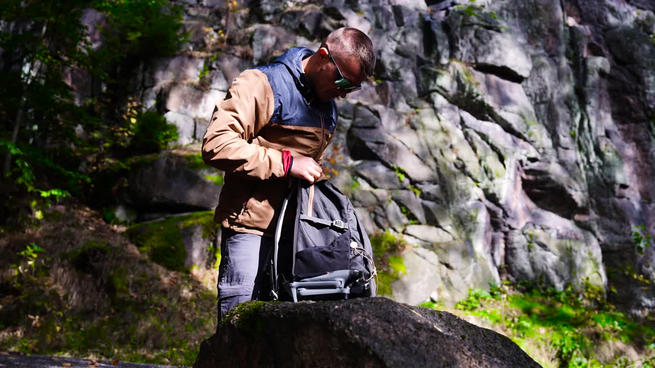 Male remove compact drone from backpack side pocket, cliff in background