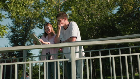 lady shows boy something from her tablet as both smile while he points at tablet with hand tattoo, background featuring greenery and trees, capturing moment of happiness and bonding