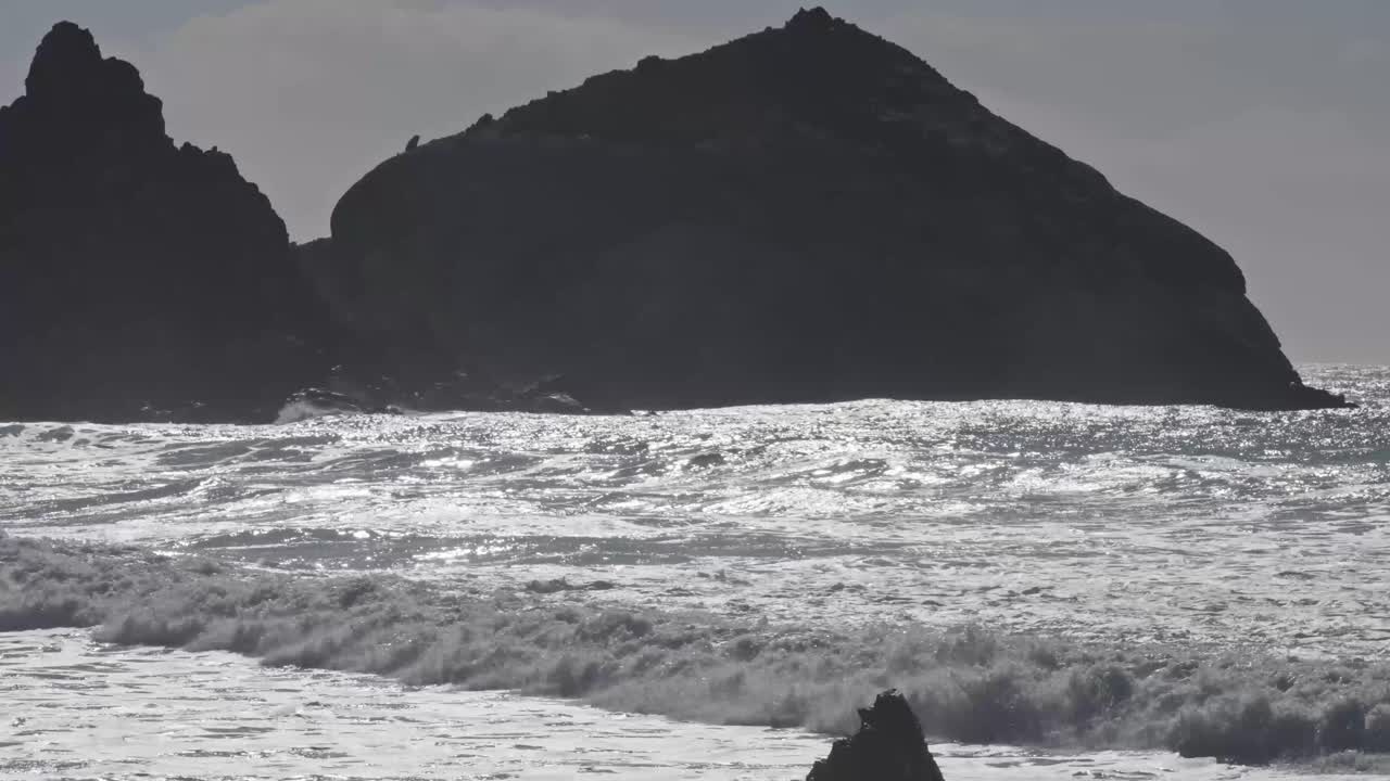Powerful waves crash against a large rock formation silhouetted by the evening sun on a rugged Pacific shoreline.