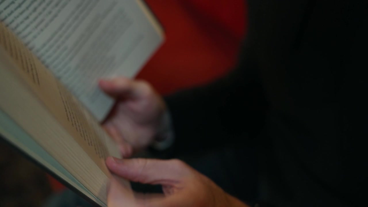Close-up of a hand reading an open book indoors, with soft focus pages, warm background color and calm literature lifestyle atmosphere.
