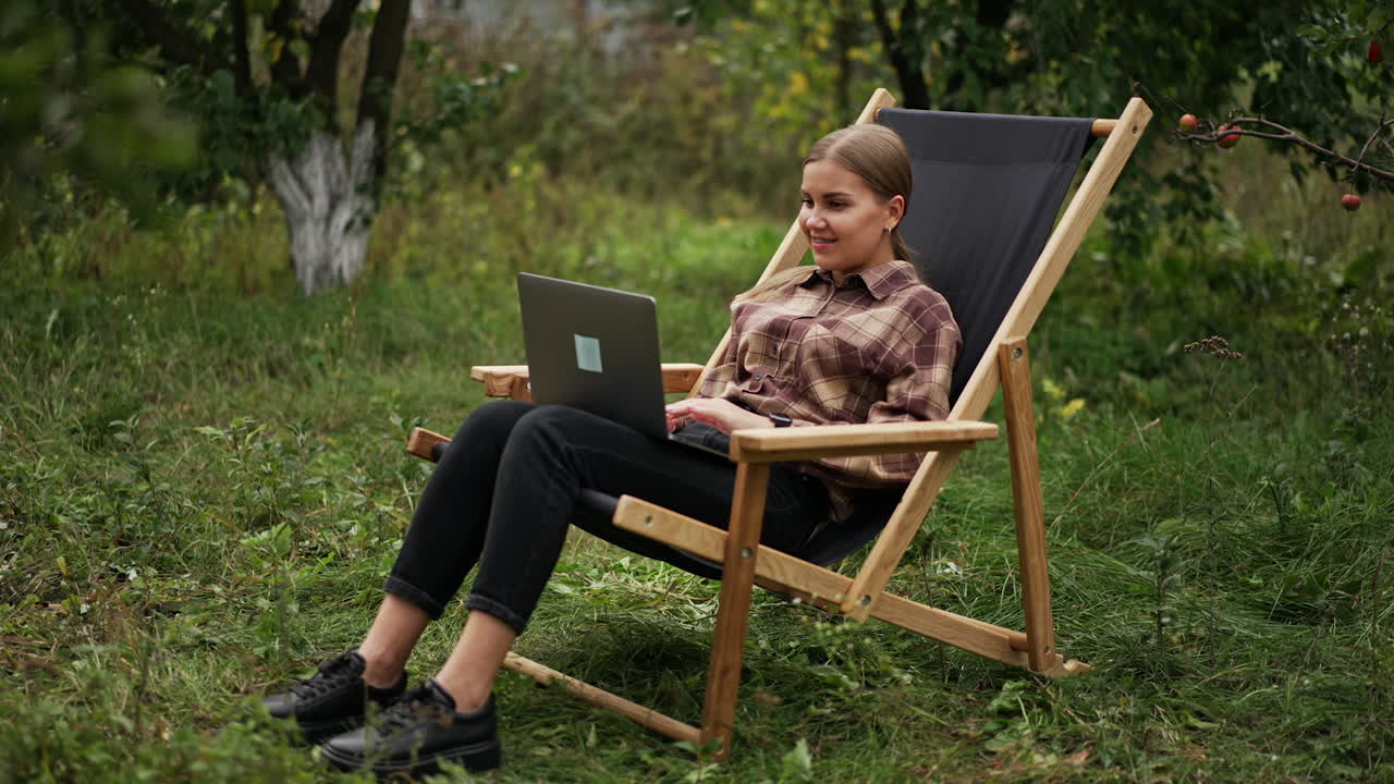 Beautiful woman in chequered shirt starts work on her computer sitting in garden chair. Positive relaxed lady opens laptop and begins typing. Garden at backdrop.