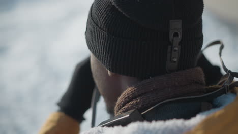 back view of cameraman wearing knit beanie and scarf, gloved hands adjusting camera lens while walking snowy sidewalk under winter sun, capturing blurred urban buildings and distant traffic