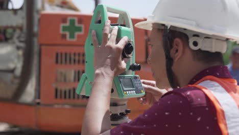 joven ingeniero con casco de seguridad y chaleco reflectante en un sitio de construcción midiendo y examinando la ubicación con una excavadora en el fondo en filipinas