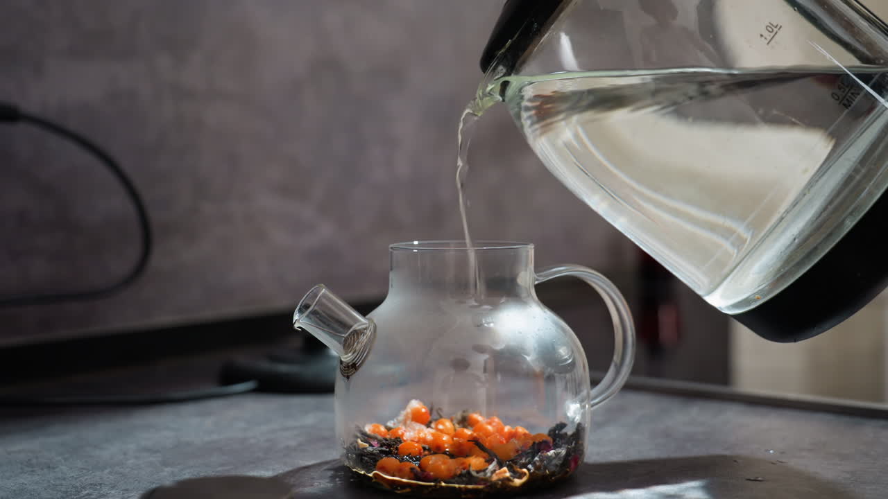 Teiera di vetro piena di acqua calda, bacche d'arancia essiccate che si depositano sul fondo, flusso trasparente del bollitore e minuscolo schizzo sul piano di lavoro, vapore aromatico che sale, primo piano di una scena in cucina che cattura la lenta infusione