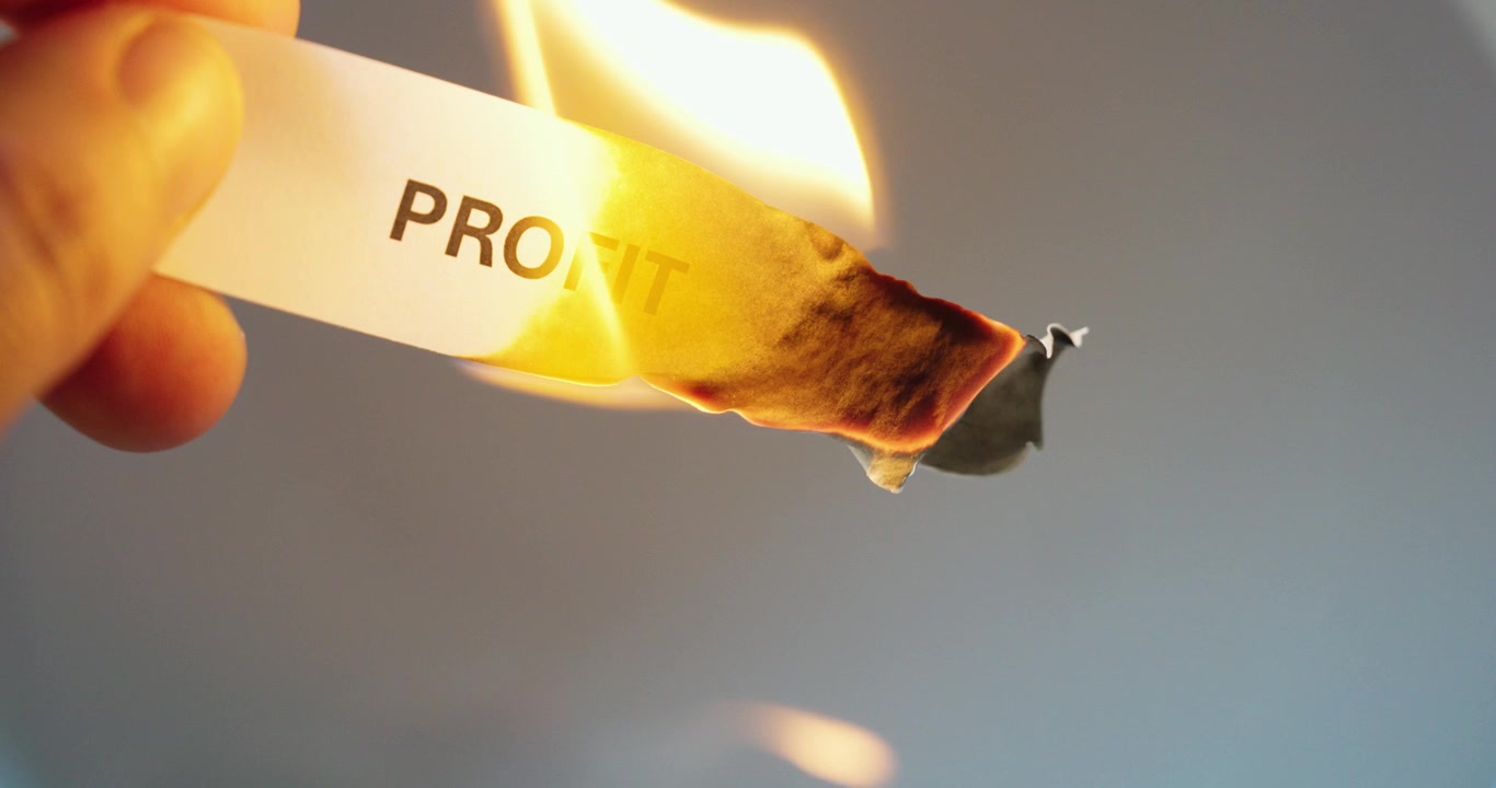 Primo piano di una striscia di carta con la parola “profitto” che brucia, a simboleggiare la perdita finanziaria, il rischio aziendale, la pressione del mercato, la crisi economica e il calo dei ricavi