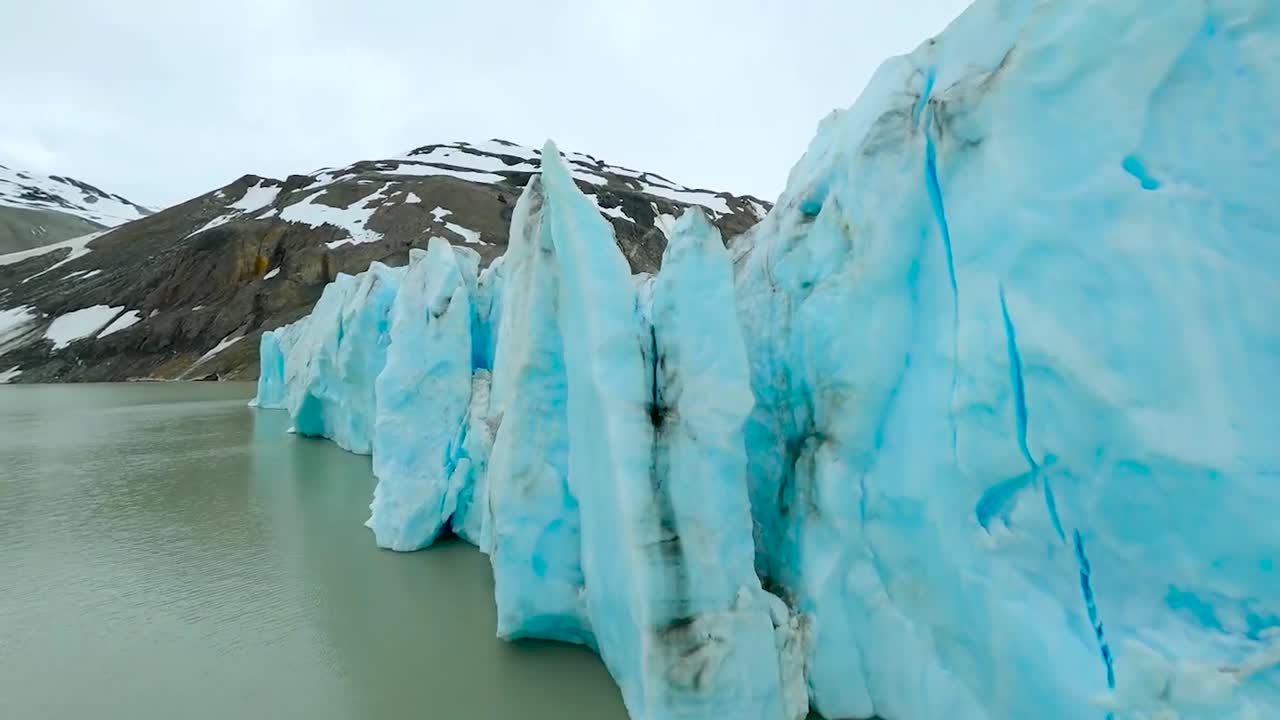 FPV aerial drone gliding over, through and backwards between icy blue and tall steep glaciers in Greenland or Iceland during a cloudy day while a big brown snow covered mountain is visible in the back