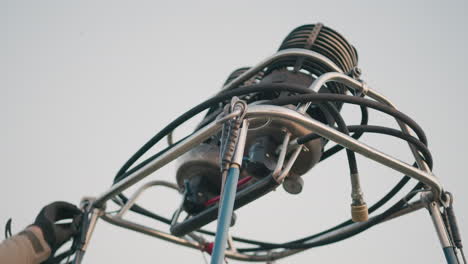 close up of hot air balloon burner assembly showing metal frame structure, braided hoses, safety cables, pressure gauge, valves and coils against clear sky