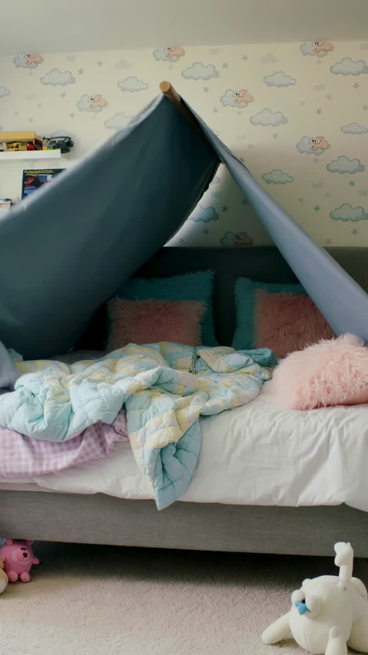 Vertical video: Bringing pink pillow girl entering right, kids building cozy fort on bed under tent