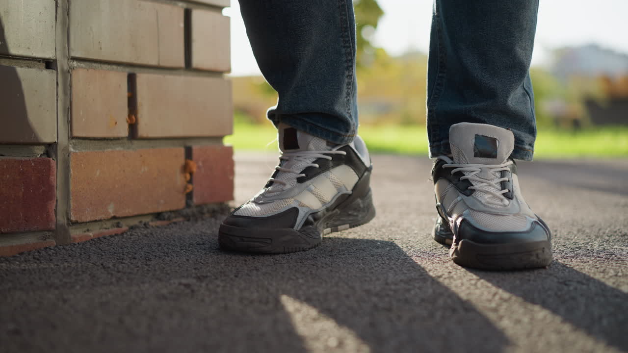 leg view of man in jeans and sneakers gently tapping right foot on asphalt surface near brick wall with shadow cast on ground under sunlight and blurred greenery in background