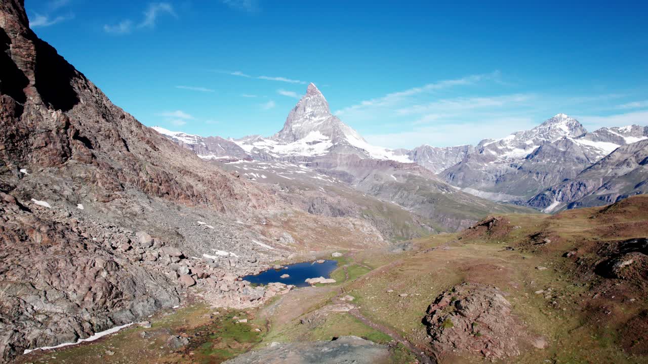 L'iconica vetta del Cervino che si erge sopra una valle alpina, terreno roccioso e piccoli laghi di montagna sotto un cielo azzurro e limpido, mostrando un classico paesaggio delle Alpi svizzere.