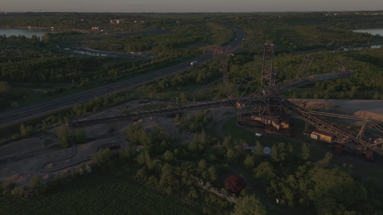 Drone orbit of a massive industrial structure near Leipzig, Germany. Captures a sunset-lit highway, surrounding forest, and lakes of the Leipzig Neuseenland in the background.