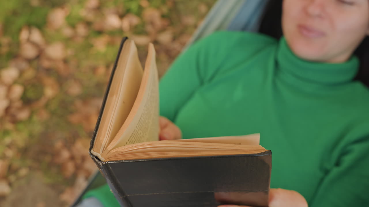 Primo piano delle pagine di un diario di una donna caucasica in campeggio. Mani che sfogliano pagine di pelle testurizzata, con l'amaca inquadrata all'aperto. Sfondo di foglie autunnali, lettura concentrata e umore riflessivo. Dettaglio di un quaderno rustico, un momento lento.