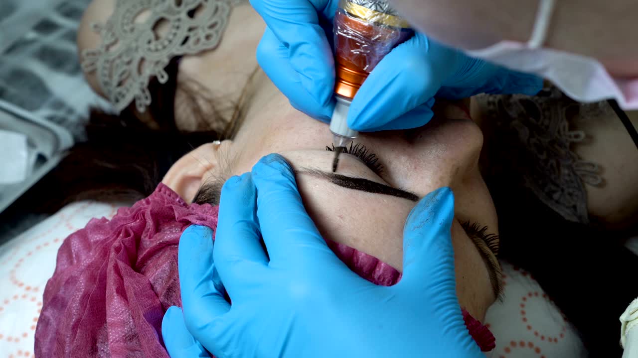 maquillaje permanente. cosmetólogo lleva a cabo el procedimiento de microblading y aplica pintura negra con una aguja en las cejas de una chica en un salón de belleza. muy de cerca. 4k. 25 fps.