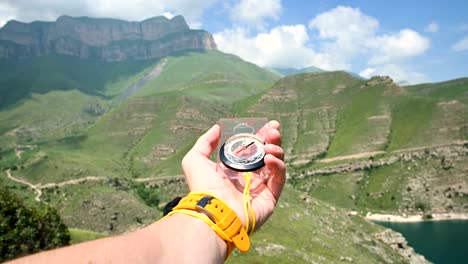 the first-person view of a male hand holding a plastic magnetic compass rotates on the side and looking for the right direction in the background of a mountain. the concept of orienteering