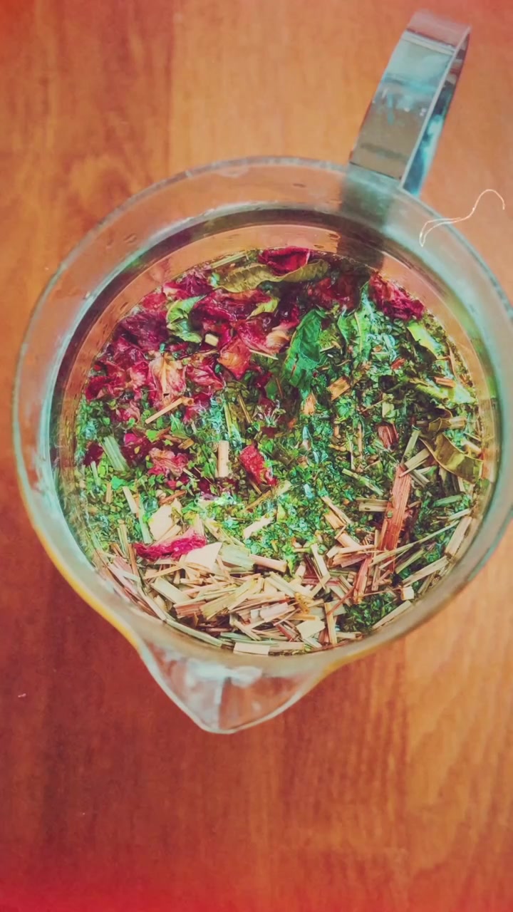 En glastekande fyldt med levende tørrede urter og blomsterblade får hældt varmt vand i, hvilket blander ingredienserne i et roligt køkkenmiljø