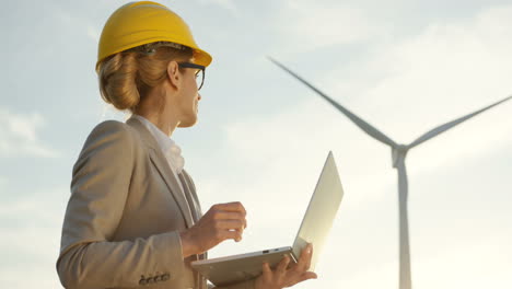 vista inferior de una ingeniera caucásica que usa un casco usando una computadora portátil mientras verifica el funcionamiento de las turbinas del molino de viento