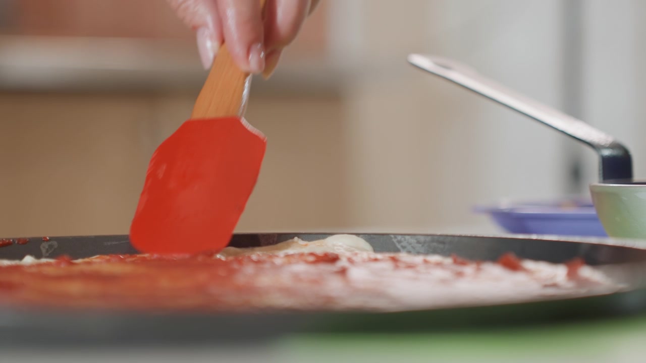 Vista cercana en ángulo bajo de una mano usando una espátula de silicona roja para untar salsa brillante sobre masa plana o repostería, mostrando una aplicación suave, brillo húmedo, vetas texturizadas y una ligera acumulación en el borde.