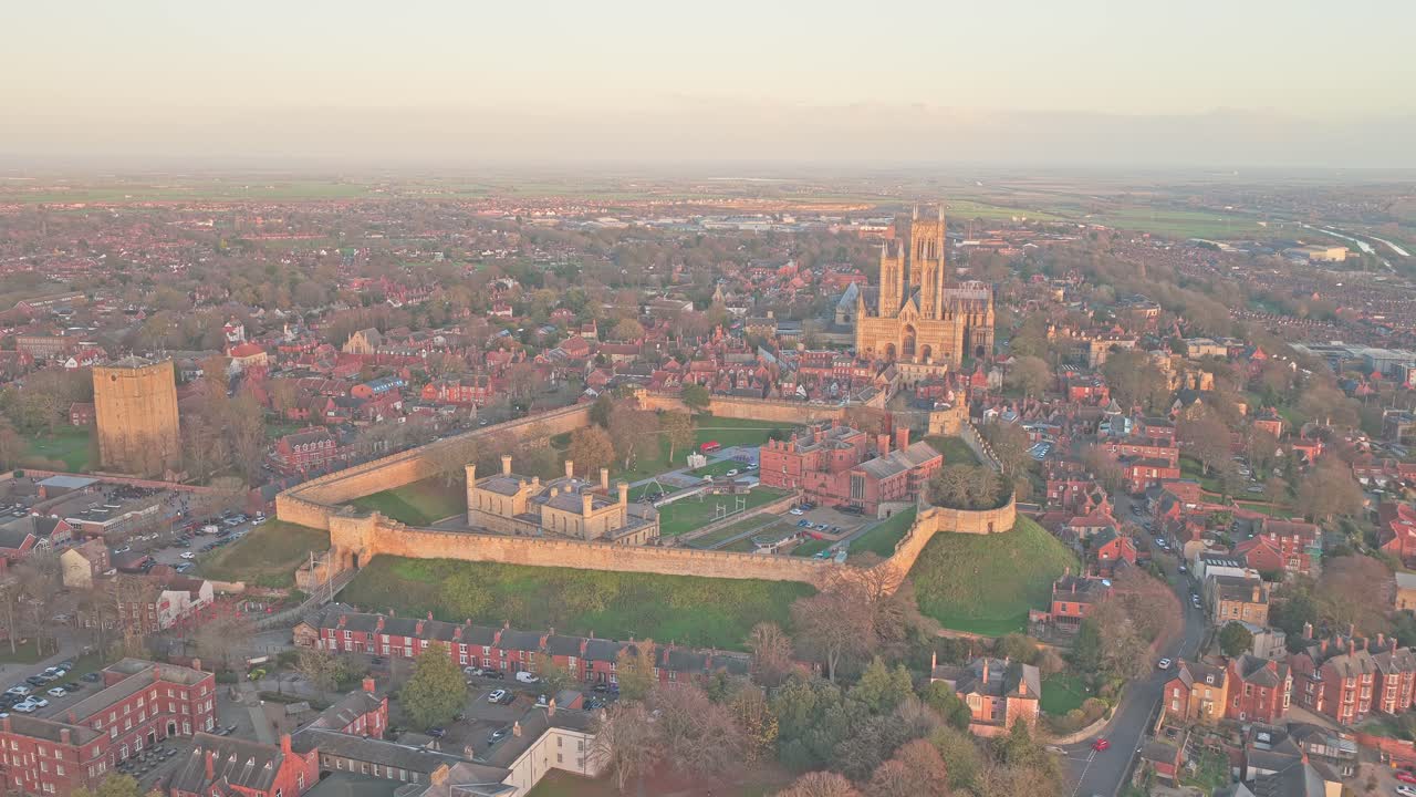 Lincoln Cathedral and Lincoln Castle dominating the historic city skyline, illuminated by the warm light of sunrise or sunset, presenting a panoramic aerial view of the urban landscape