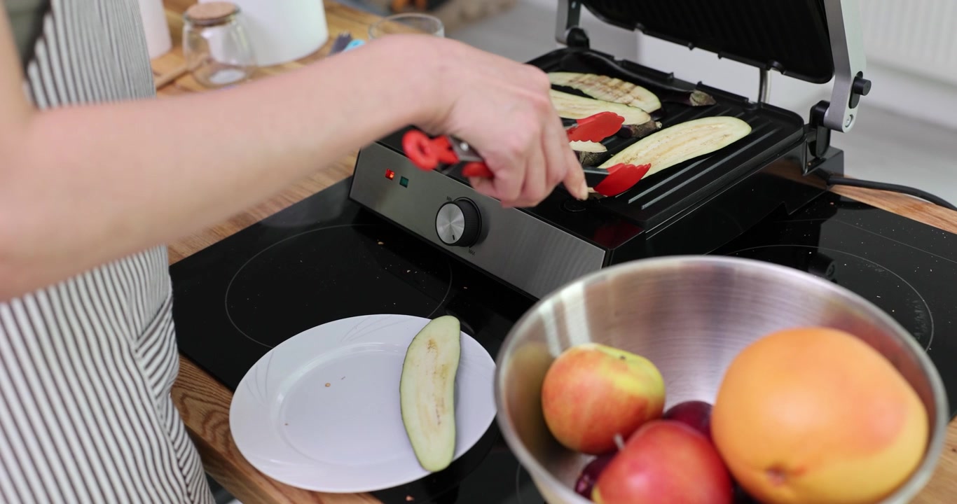 Female chef in apron grills eggplant on electric device