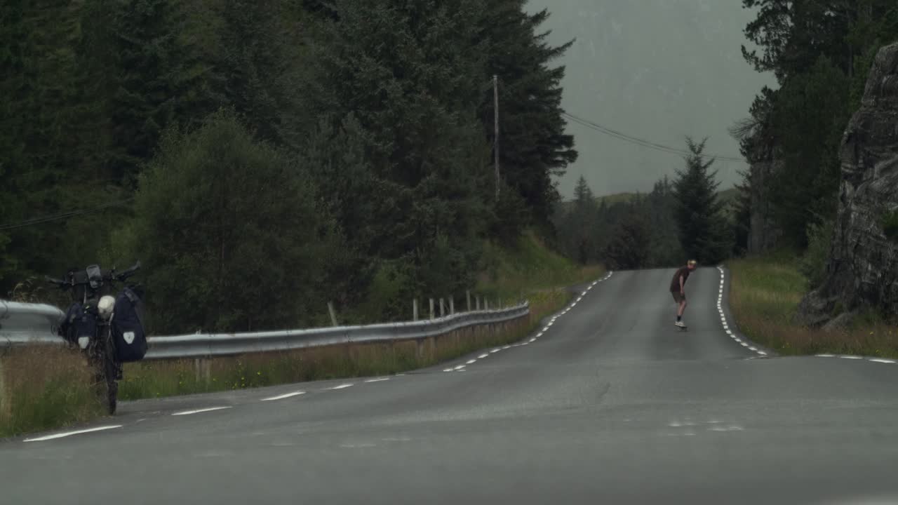 악천후를 배경으로 한 황량한 도로에서 카메라에서 멀리 떨어진 곳에서 묘기를 부리며 자전거 여행에서 휴식을 취하는 스케이트보더