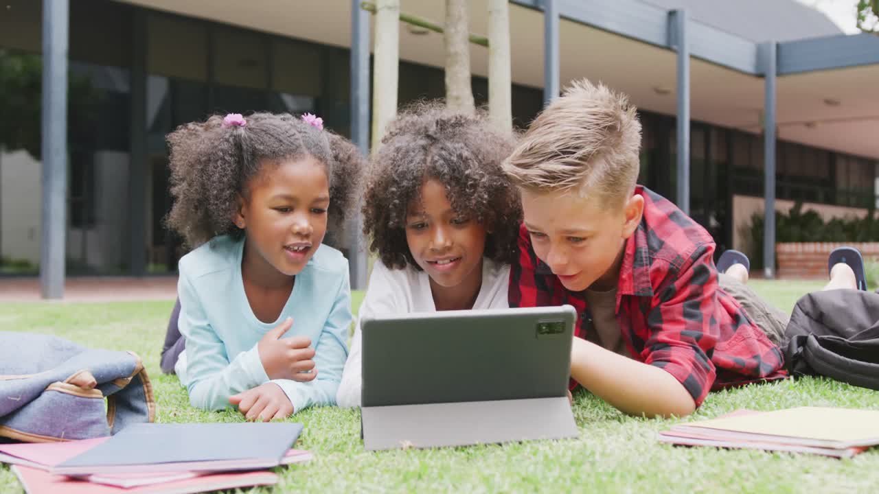 video de tres escolares felices y diversos viendo una tableta tirada en el patio de la escuela, espacio de copia