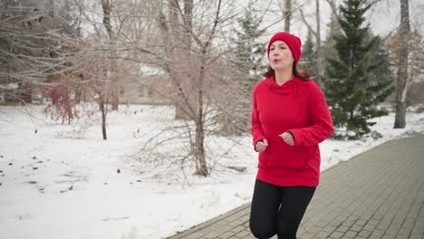 tired lady in red beanie and hoodie jogging outdoors during winter surrounded by snow-covered trees, serene park pathway, evergreen pines, foggy atmosphere, and residential building in background