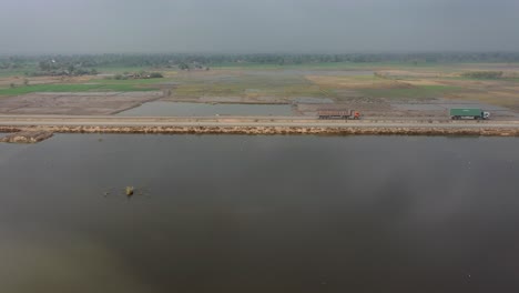 Aerial-Over-River-With-View-Of-Lorries-Going-Past-On-Highway-With-Farmland-In-the-Distance