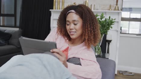 retrato de una mujer biracial feliz relajándose en el sofá usando una tableta en casa, sonriendo, en cámara lenta.