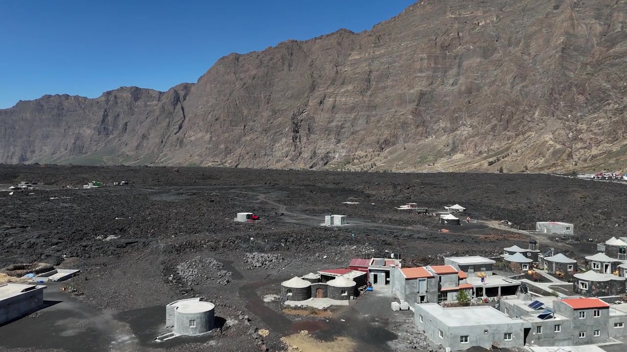 Luftfoto med drone af landsbyen Chã das Caldeiras og traditionelle stenhuse beliggende på sort vulkansk jord under den massive Bordeira klippevæg i Fogo Nationalpark, Kap Verde.