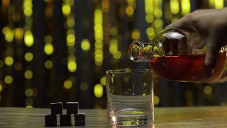 pouring of golden whiskey, cognac or brandy from bottle into glass with rocks ice stones on table