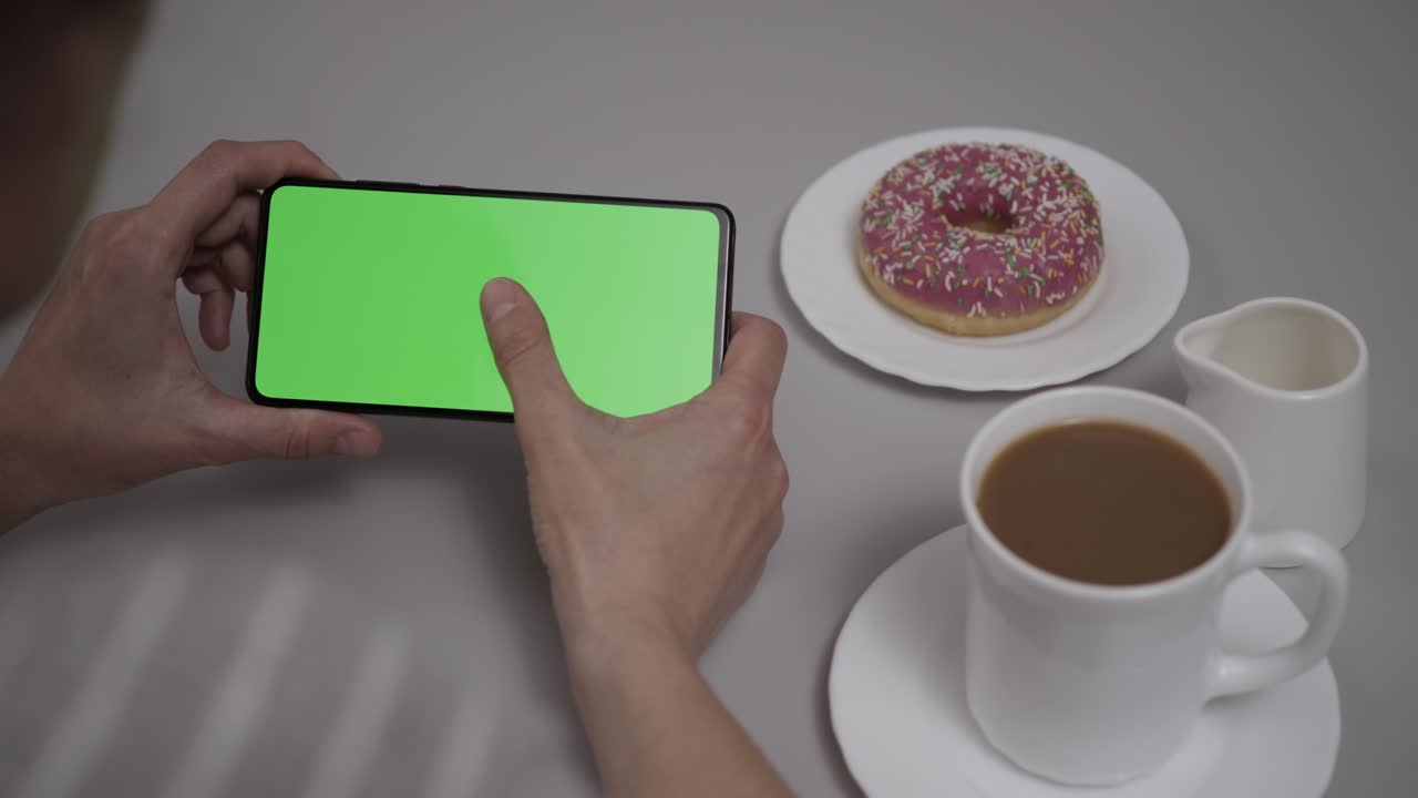 mujer sentada en la mesa de café donut usando un teléfono inteligente con pantalla verde de clave de croma, desplazándose a través de las redes sociales medios de comunicación en línea tienda internet. teléfono inteligente en modo horizontal con maqueta de pantalla verde.