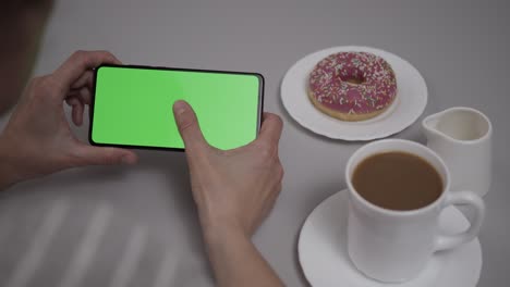 woman sitting at table coffee donut using smartphone with chroma key green screen, scrolling through social network media online shop internet. smartphone in horizontal mode with green screen mock-up.