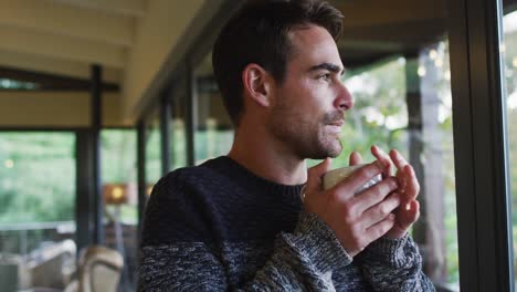 Thoughtful-caucasian-man-looking-out-of-window-and-drinking-coffee-in-dining-room