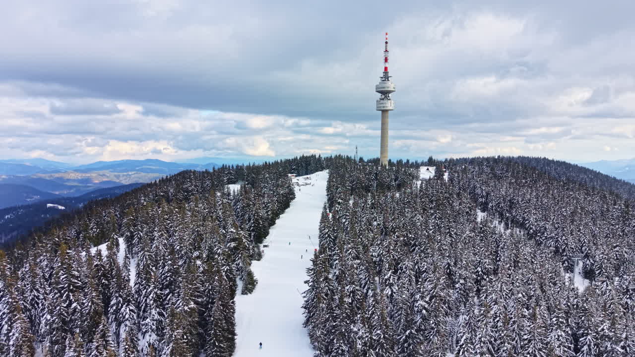 Montagna innevata con persone che sciano e un'alta torre sullo sfondo in una giornata nuvolosa.