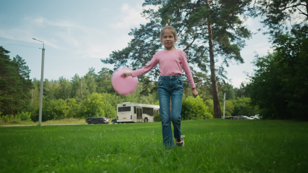 Little girl in pink shirt and blue jeans walking on grassy field holding pink balloon, playing joyfully with parked bus and cars in background under clear blue sky and tall pine trees