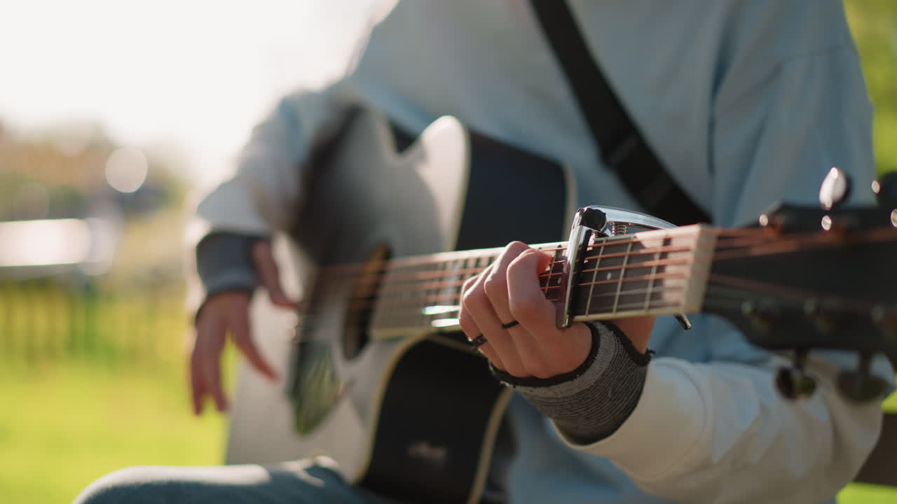 Chitarrista all'aperto che suona la chitarra acustica, primo piano delle mani sulla tastiera e sulla paletta, bokeh del parco illuminato dal sole, maglione e jeans casual, fingerpicking e strumming, sessione di pratica sulla panchina, caldo