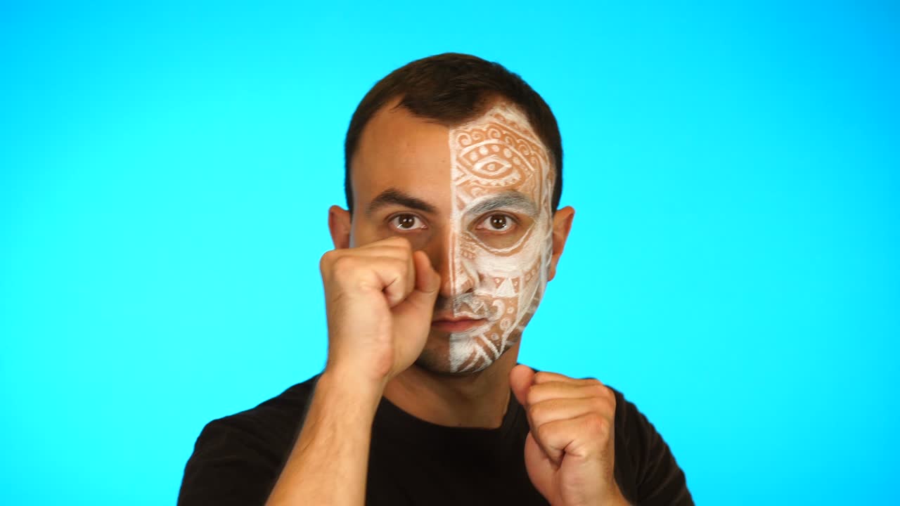 Man with painted face is standing in boxer pose and looking at camera against blue background