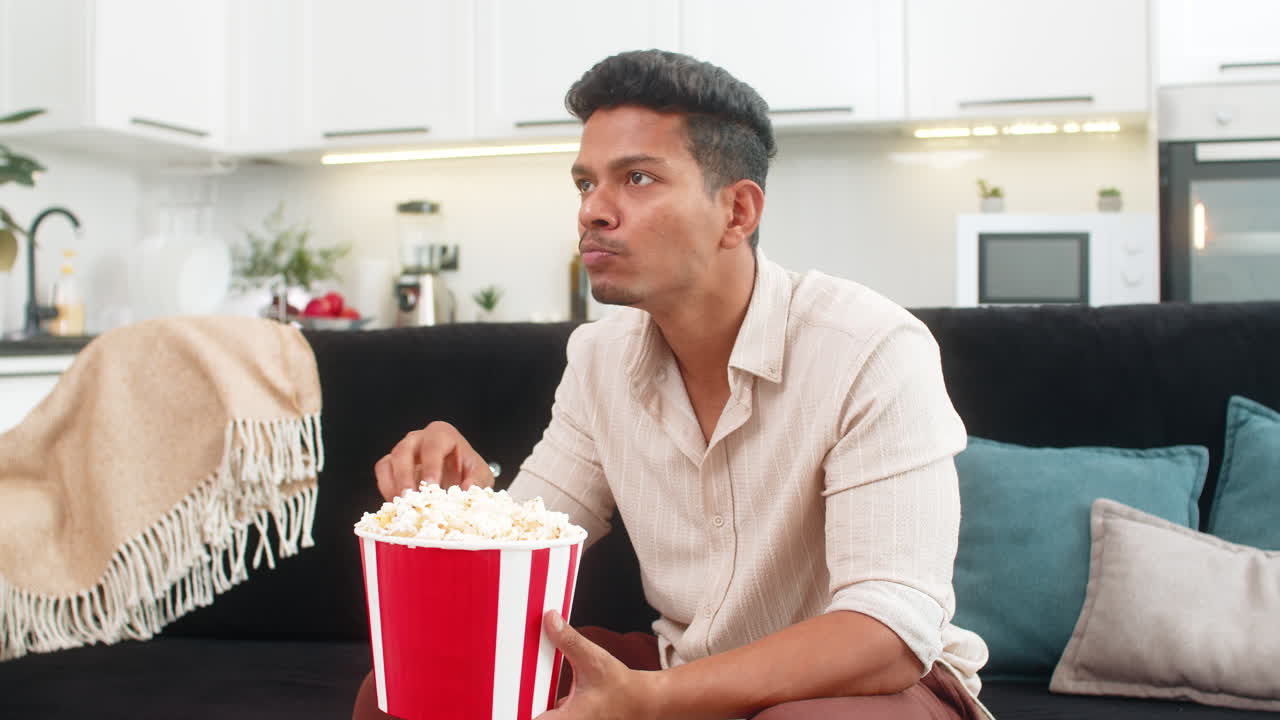 Uomo indiano guarda il trailer di un film in TV, mangia popcorn, ride e reagisce a momenti divertenti, commentando ad alta voce.