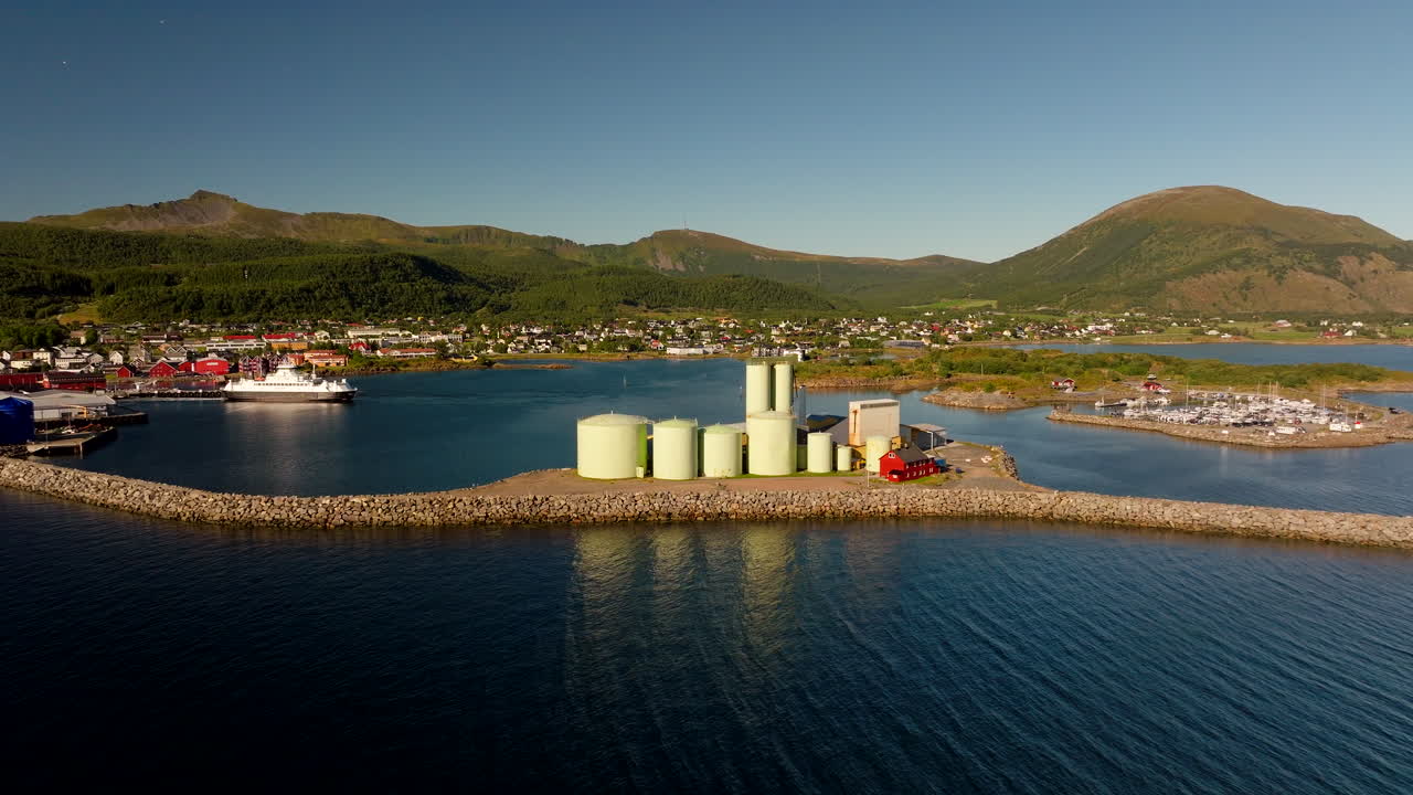 Filmatisk drönarrörelse framåt över fiskerisilos vid Melbu hamn, Hadsel, Lofoten, Norge. Kuststad och bergskuliss ovan lugnt vatten under klar himmel