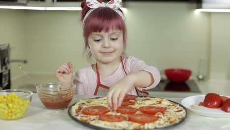 cooking pizza. little child in apron adding sliced tomatoes to dough in kitchen